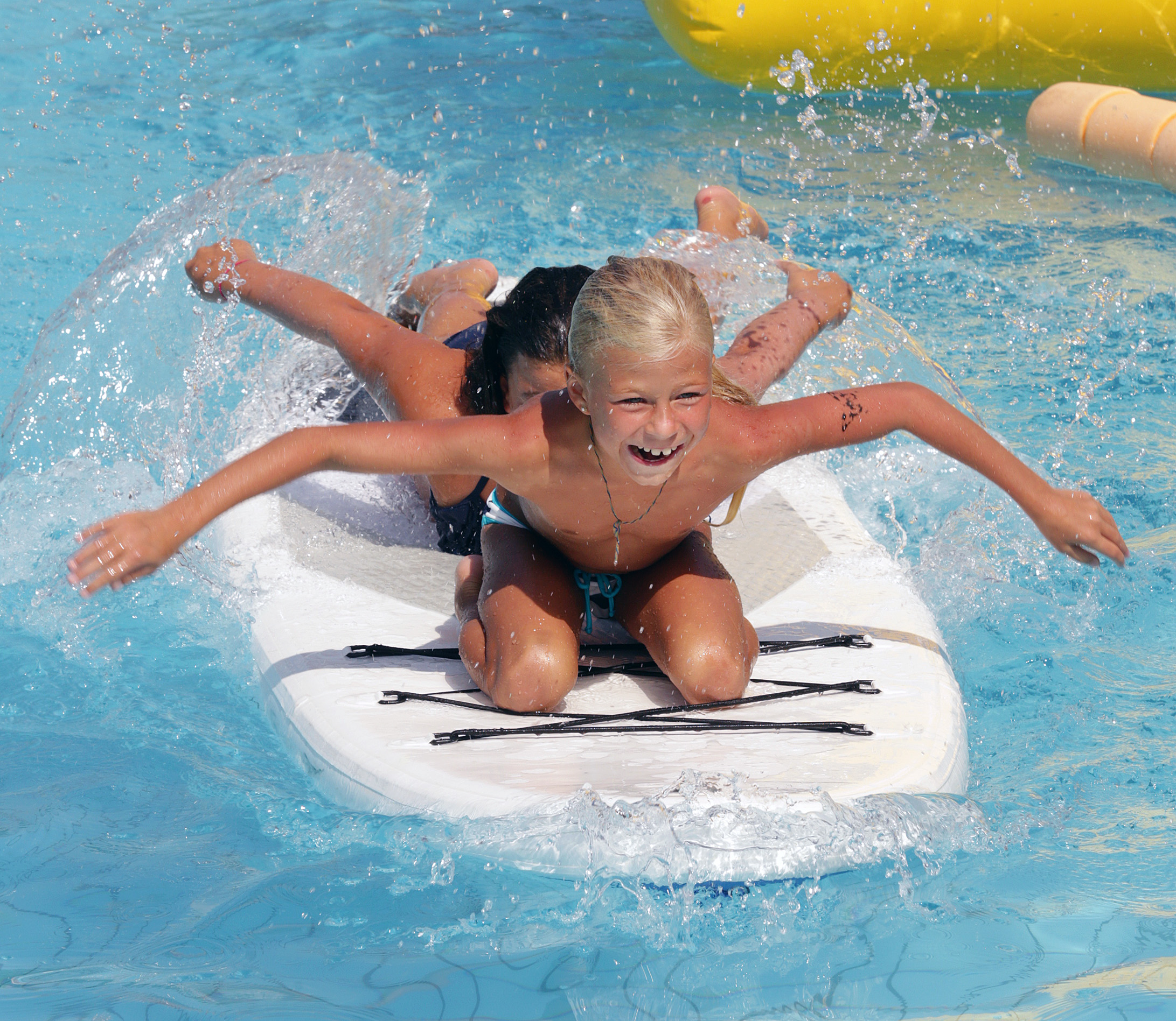 Zwei Kinder rutschen lachend auf einem weißen Surfbrett im Schwimmbecken, Wasser spritzt um sie herum