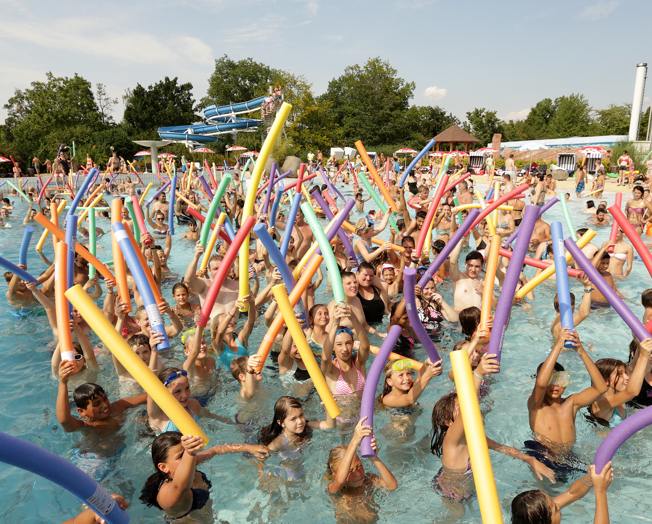 Viele Menschen unterschiedlichen Alters im Schwimmbecken eines Freibads halten bunte Schwimmnudeln in die Luft, im Hintergrund eine blaue Wasserrutsche und Bäume