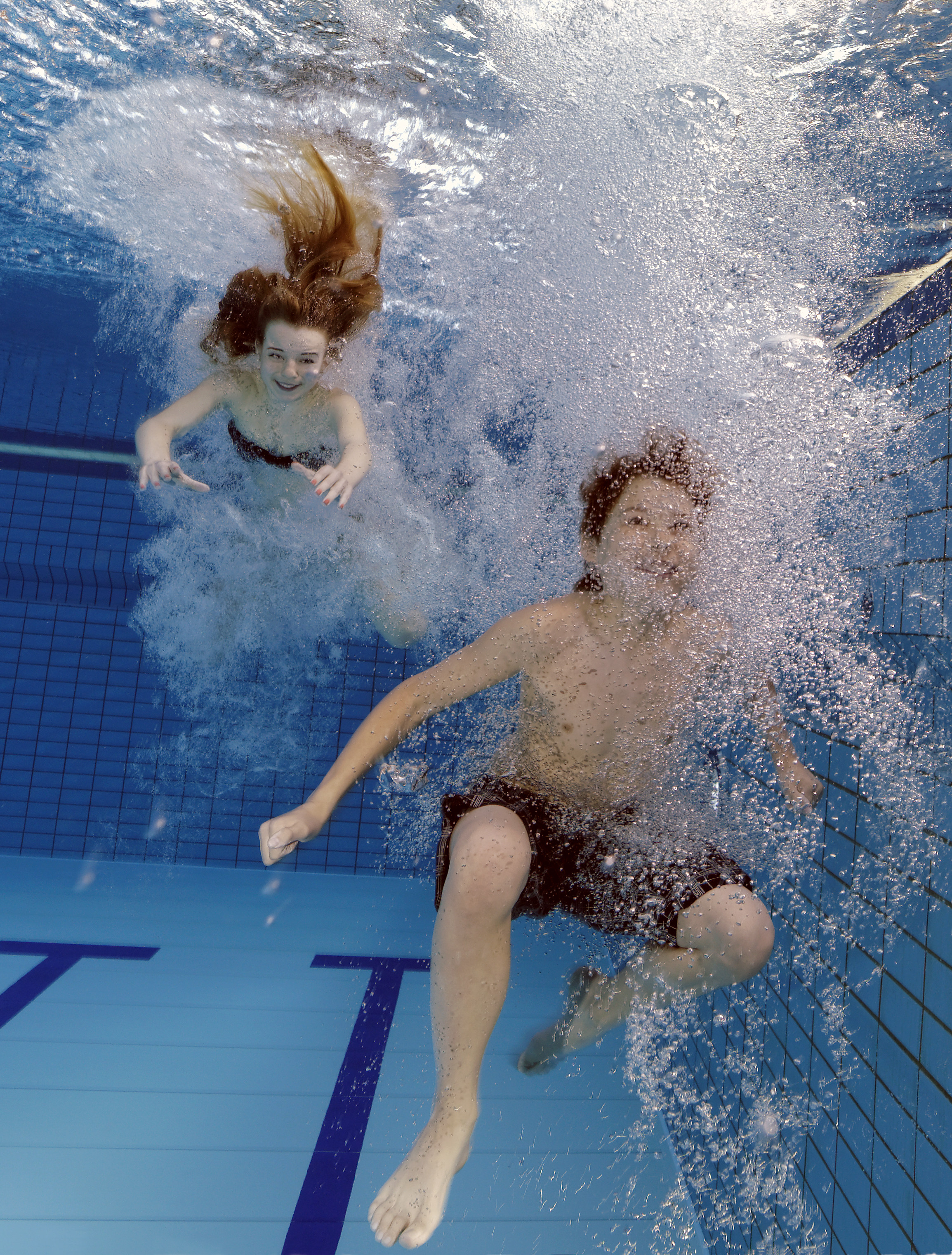 Zwei Kinder tauchen unter Wasser in einem Schwimmbecken mit blauen Kacheln und schwarzen Linien auf dem Beckenboden
