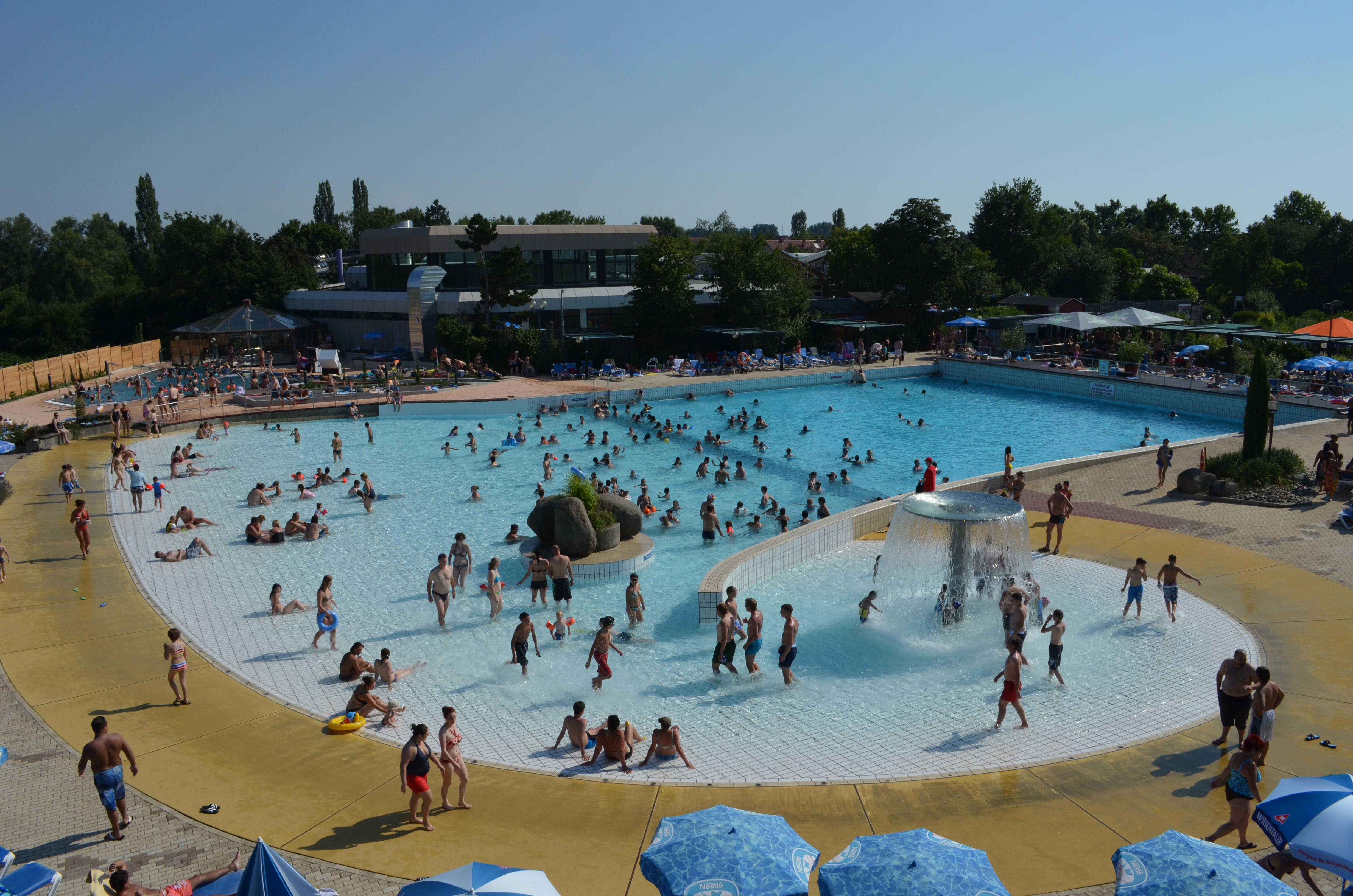 Belebtes Freibad mit vielen Menschen im Wasser und auf der Liegewiese an einem sonnigen Tag
