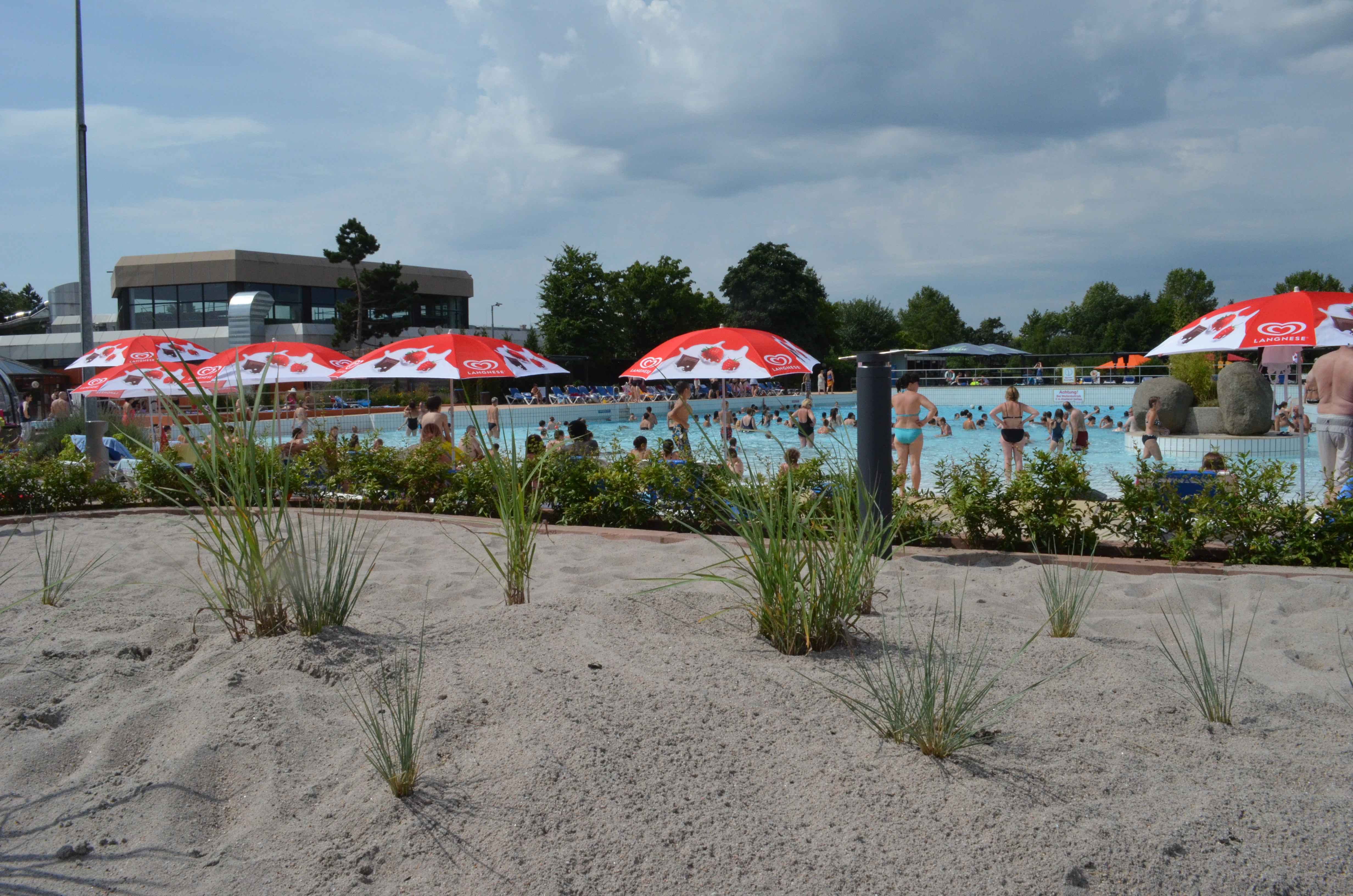 Freizeitbad mit Sandstrand, roten Sonnenschirmen und vielen Menschen im Schwimmbecken bei bewölktem Himmel