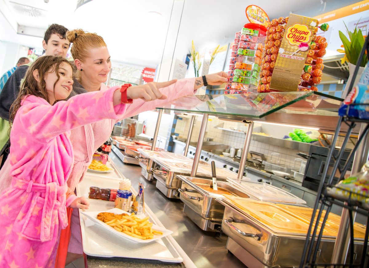 Drei Personen in Bademänteln wählen Essen an einem Buffet mit warmen Speisen und Süßigkeiten im Hintergrund