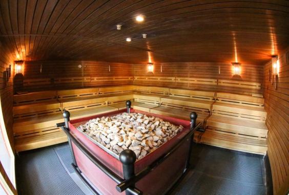 Innenraum einer Sauna mit Holzwänden und Holzbänken, zentraler Ofen mit Steinen umgeben von einem Metallgitter
